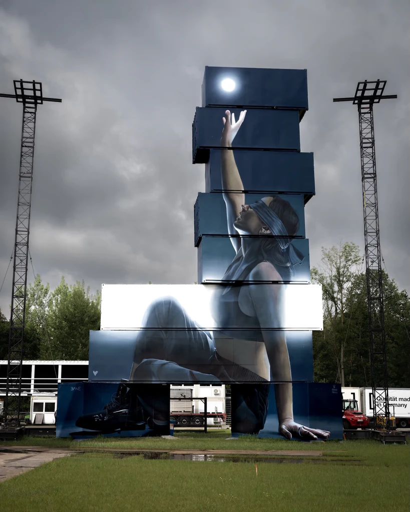Grosses Mural auf gestapelten Containern zeigt eine Frau, die nach einer leuchtenden Kugel greift, vor bewölktem Himmel in Belgien.